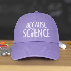 May include: A lavender baseball cap with the words "BECAUSE SCIENCE" embroidered in white. The design includes a beaker graphic. The cap is made of a soft material and has a curved brim.