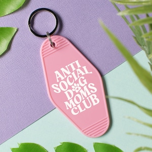 May include: Pink motel key chain with a white inscription that reads "Anti Social Dog Moms Club".