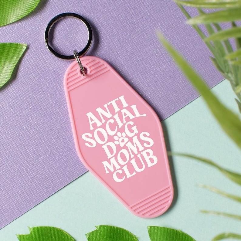 May include: Pink motel key chain with a white inscription that reads "Anti Social Dog Moms Club".