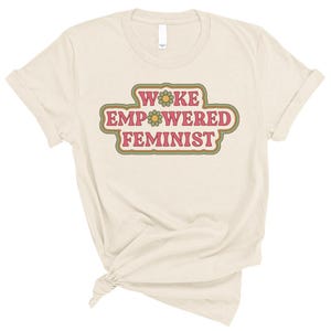 May include: Cream-colored t-shirt with the words "WOKE EMPOWERED FEMINIST" in a retro, layered design. The text is in pink, green, and orange, with flower accents. The shirt is made of soft material and has short sleeves.