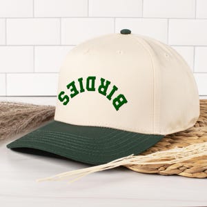 May include: A cream-colored baseball cap with a dark green brim and button. The word "BIRDIES" is arched across the front in green lettering. The hat is sitting on a woven surface.