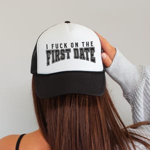 May include: A black and white trucker hat with the text "I FUCK ON THE FIRST DATE" in bold, black letters. The hat has a white front panel and a black brim and mesh back.