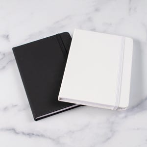 K&ouml;nnte beinhalten: Zwei rechteckige Notizb&uuml;cher, eines schwarz und eines wei&szlig;, werden auf einer Marmoroberfl&auml;che gezeigt. Jedes Notizbuch hat einen passenden Gummibandverschluss. Das wei&szlig;e Notizbuch liegt auf dem schwarzen.