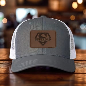 May include: A gray trucker hat with a white mesh back and a brown leather patch. The patch features a stylized "S" logo and the word "SUPERDAD" in a bold font. The hat is displayed on a wooden surface.