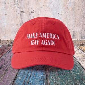 May include: Red baseball cap with white embroidered text that reads "Make America Gay Again".