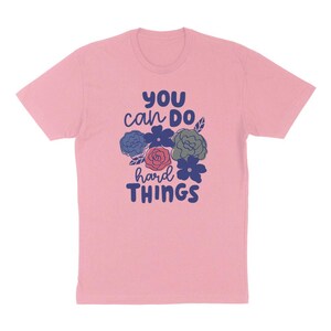 Puede incluir: Camiseta rosa claro con el texto "YOU can DO hard THINGS" en azul y blanco. El dise&ntilde;o incluye ilustraciones florales en azul, rojo y verde.
