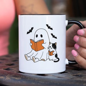 May include: White ceramic mug with a black handle and rim. The mug features a cartoon ghost and a cat, both reading orange books, with black bats flying above. A fun, Halloween-themed design.