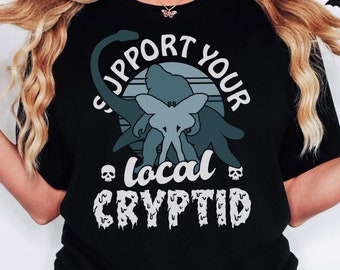 May include: Black t-shirt with a graphic design featuring a Bigfoot, Mothman, and a dinosaur with the text "Support Your Local Cryptid".