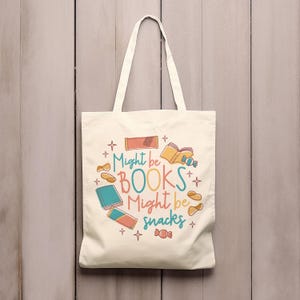 May include: A white canvas tote bag with a colourful design featuring the text "Might be BOOKS Might be snacks" and illustrations of books, snacks, and stars.