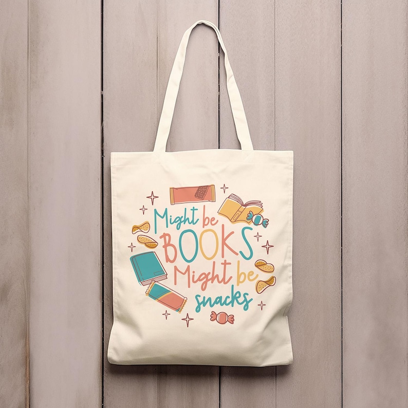 May include: A white canvas tote bag with a colourful design featuring the text "Might be BOOKS Might be snacks" and illustrations of books, snacks, and stars.