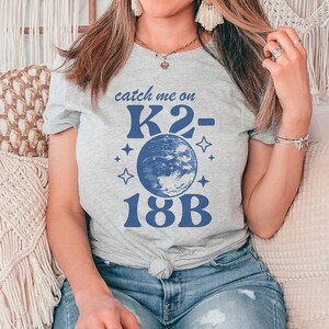 May include: A gray t-shirt with a blue graphic that says "catch me on K2-18B". The graphic also includes a blue image of the Earth and stars.