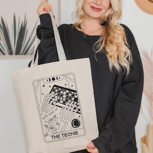 May include: Beige tote bag with a black and white graphic of skeleton hands typing on a keyboard, surrounded by stars, moons, and roses. Text on the bag reads "NEVER LOGGED OFF" and "THE TECHIE". The bag has gray straps.