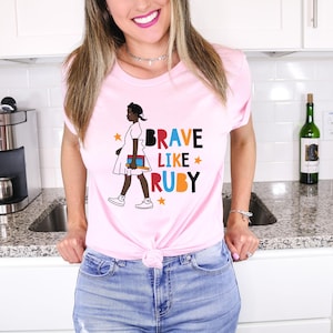 May include: Pink t-shirt with a graphic of a young girl walking and holding a book. The text "BRAVE LIKE RUBY" is printed in colorful letters on the shirt.