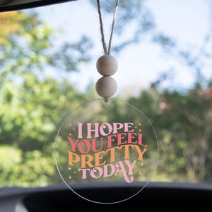 May include: A clear, circular car air freshener with the text "I HOPE YOU FEEL PRETTY TODAY" in pink, orange, and red. The air freshener hangs from a string with two wooden beads. The background is a blurred view of green foliage.