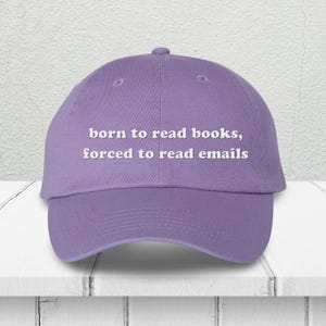 May include: A lavender baseball cap with the text "born to read books, forced to read emails" in white. The cap is made of a soft material and has a curved brim. The hat is sitting on a white surface.