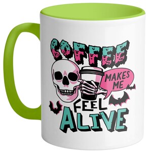 May include: White ceramic coffee mug with a green handle and interior. The mug features a skull graphic holding a coffee cup, with the text "COFFEE MAKES ME FEEL ALIVE" in a spooky font. Bats are also included in the design.