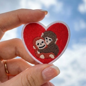 May include: Embroidered heart-shaped patch with a red border and a white outline. The patch features two hugging monkeys in brown and orange, set against a blue sky background. A cute and charming design.