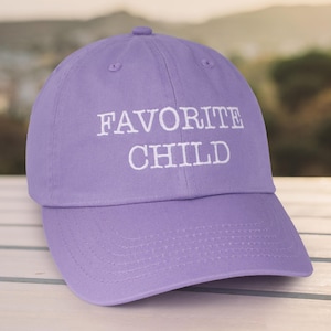 May include: A purple baseball cap with white embroidered text that reads "FAVORITE CHILD".