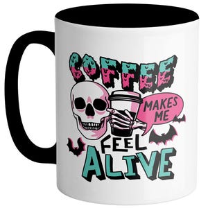 May include: White ceramic coffee mug with a black handle and interior. The mug features a skull graphic holding a coffee cup, with the words "COFFEE MAKES ME FEEL ALIVE" in a teal, pink, and black design.