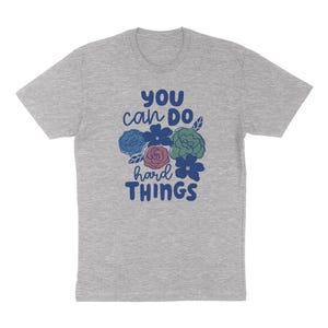 Puede incluir: Camiseta gris jaspeado con la frase "You can do hard things" en azul, blanco y rosa, acompa&ntilde;ada de ilustraciones florales. La camiseta tiene mangas cortas y cuello redondo. Las ilustraciones florales son azules, verdes y rosas.