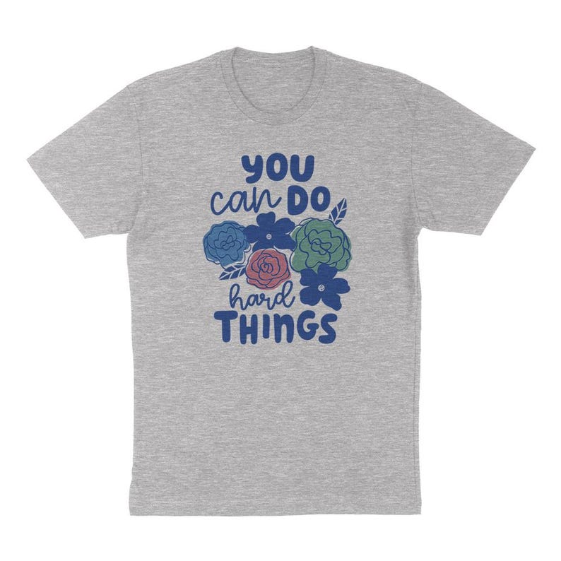 Puede incluir: Camiseta gris jaspeado con la frase "You can do hard things" en azul, blanco y rosa, acompa&ntilde;ada de ilustraciones florales. La camiseta tiene mangas cortas y cuello redondo. Las ilustraciones florales son azules, verdes y rosas.