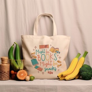 May include: A white canvas tote bag with the text "Might be BOOKS Might be snacks" in colourful letters. The bag is decorated with illustrations of books, snacks, and stars.