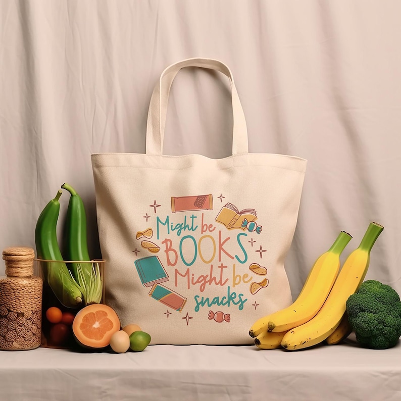 May include: A white canvas tote bag with the text "Might be BOOKS Might be snacks" in colourful letters. The bag is decorated with illustrations of books, snacks, and stars.