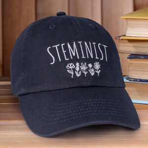 May include: A black baseball cap with the word "STEMINIST" embroidered in white on the front. The word is surrounded by a row of small white flowers.