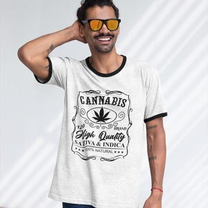 May include: A light gray t-shirt with black trim around the collar and sleeves. The shirt features a black and white graphic that reads "Cannabis" with a cannabis leaf and the words "High Quality Sativa & Indica 100% Natural."