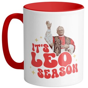 May include: White ceramic coffee mug with a red rim. The mug features a retro-style graphic of a man in a red robe with the text "It's Leo Season".