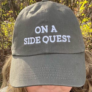May include: An olive green baseball cap featuring the white embroidered text ON A SIDE QUEST centered on the front panel. The cap has a structured crown, a curved brim with visible stitching, and a button at the top center of the dome.