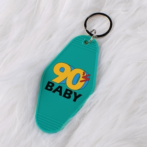May include: Teal motel-style keychain with the words "90s BABY" in a retro font. The "90" is yellow with a blue outline and a red lightning bolt. The keychain has a silver key ring.