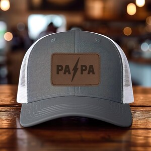 May include: A gray trucker hat with a white mesh back. The front features a brown leather patch with the word "PA PA" and a lightning bolt graphic.