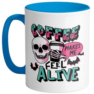 May include: White ceramic coffee mug with a blue handle and interior. The mug features a skull graphic holding a coffee cup, with the words "COFFEE MAKES ME FEEL ALIVE" in a spooky font, and bat illustrations.
