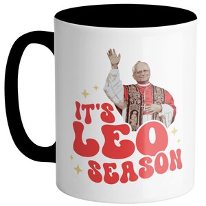 May include: White ceramic mug with a black rim and handle. The mug features a retro-style graphic of a man in a red robe with the text "It's Leo Season".