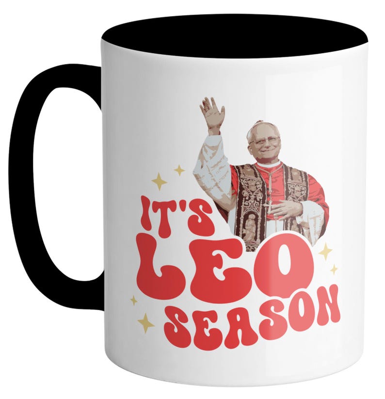 May include: White ceramic mug with a black rim and handle. The mug features a retro-style graphic of a man in a red robe with the text "It's Leo Season".