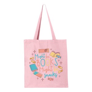 May include: A pink canvas tote bag featuring the text "Might be books might be snacks" in a colourful font. The text is surrounded by illustrations of books, snacks, and stars.