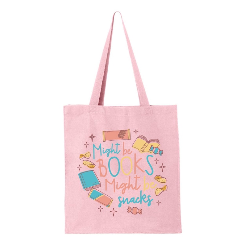 May include: A pink canvas tote bag featuring the text "Might be books might be snacks" in a colourful font. The text is surrounded by illustrations of books, snacks, and stars.