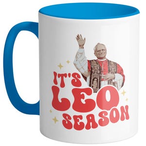 May include: White ceramic mug with a blue rim and handle. The mug features a retro-style graphic of a man in a red robe with the text "It's Leo Season".