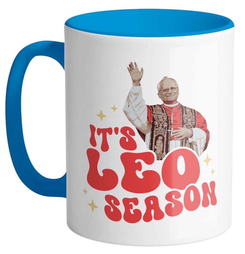 May include: White ceramic mug with a blue rim and handle. The mug features a retro-style graphic of a man in a red robe with the text "It's Leo Season".