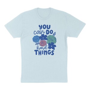 Puede incluir: Camiseta azul claro con el texto "YOU can DO hard THINGS" en azul. El dise&ntilde;o incluye un grupo de flores coloridas en azul, rosa y verde. La camiseta est&aacute; hecha de un material suave.