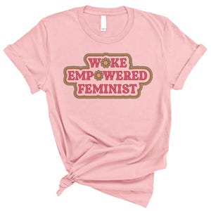 May include: A light pink t-shirt with rolled-up sleeves. The shirt features the text "WOKE EMPOWERED FEMINIST" in a retro-style design with brown and orange accents. The shirt is knotted at the bottom.