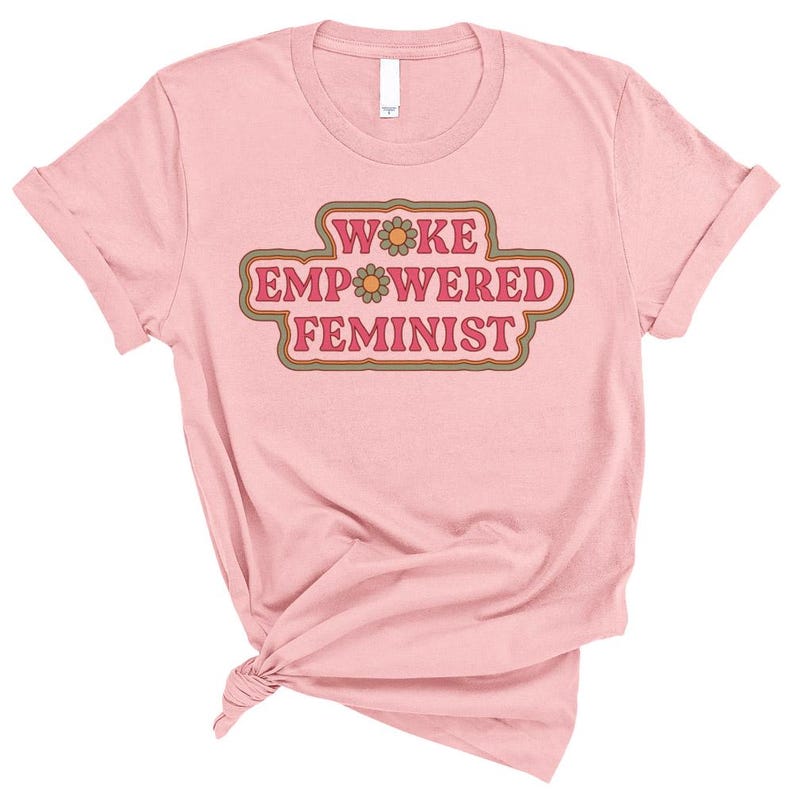 May include: A light pink t-shirt with rolled-up sleeves. The shirt features the text "WOKE EMPOWERED FEMINIST" in a retro-style design with brown and orange accents. The shirt is knotted at the bottom.