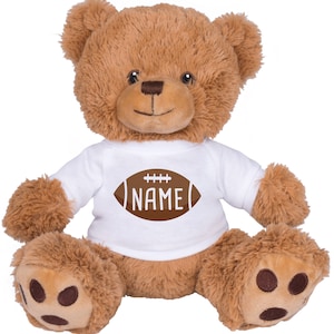 May include: A brown teddy bear wearing a white t-shirt with a brown football graphic and the word "NAME" in white. The bear has a friendly expression and is sitting with its legs outstretched.