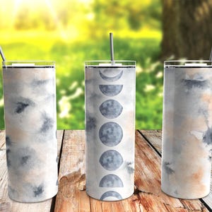 May include: Three stainless steel tumblers with clear lids and straws. The tumblers have a watercolor design in shades of gray, peach, and white. One tumbler features a moon phase design. The tumblers are displayed on a wooden surface.