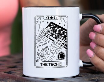 May include: White ceramic mug with a black interior and handle. The mug features a tarot card-style design with a skeleton typing on a keyboard, roses, and the text "THE TECHIE" and "NEVER LOGGED OFF".