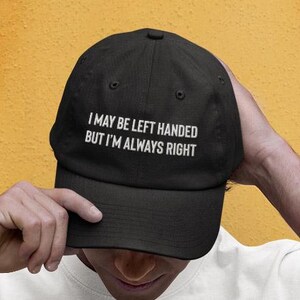 May include: A black baseball cap with white embroidered text that reads "I may be left handed but I'm always right".