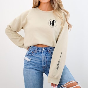 Könnte beinhalten: Beiges Langarm-Sweatshirt mit einem kreisförmigen Grafikdesign mit "HP" und Brille. Der rechte Ärmel hat ein Zauberstab-Design mit Text. Das Sweatshirt wird mit einer blauen Jeans getragen.