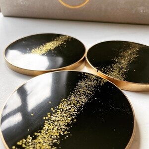 May include: Three black coasters with gold glitter accents and a gold rim. The coasters are round and have a glossy finish.