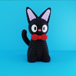 Puede incluir: Una figura de gato negro con grandes ojos blancos, una pequeña nariz rosa y una pajarita roja. El gato tiene orejas grandes y puntiagudas con el interior de color lavanda. El gato está sentado sobre una superficie azul.